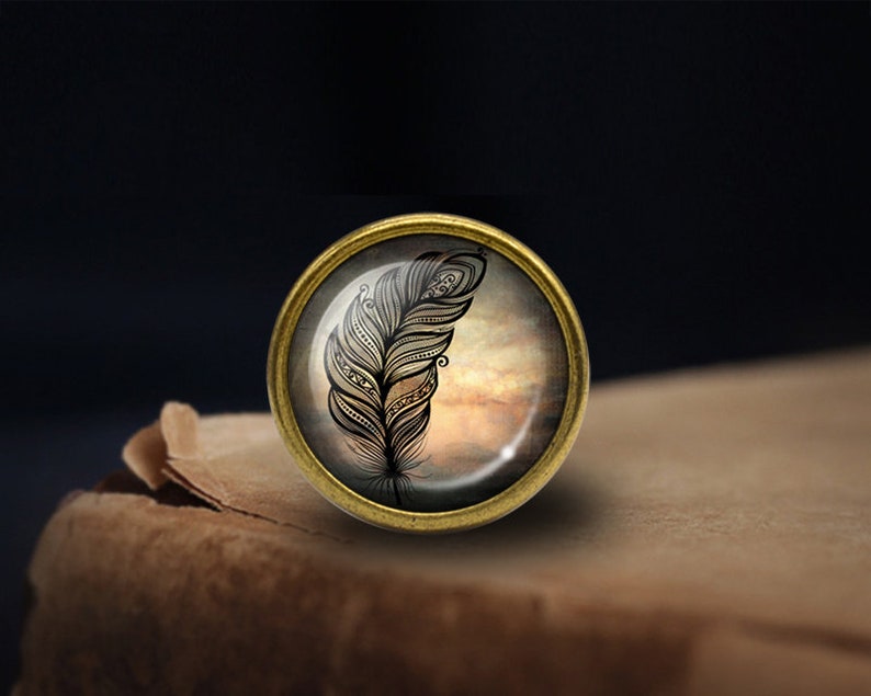 May include: A gold-toned metal pin with a glass cabochon featuring a black and white stylized feather design on a cloudy background.