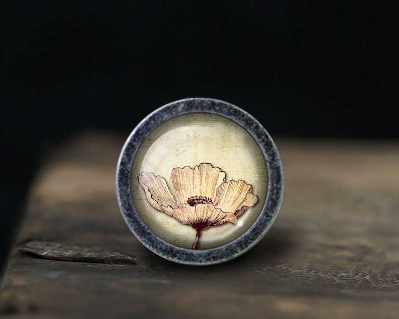 May include: A round, silver-colored knob with a floral design. The knob features a single, light brown poppy flower with a red center, set against a cream-colored background.