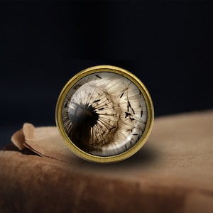 May include: A round, gold-toned pendant with a glass dome featuring a black and white image of a dandelion seed head with its seeds dispersed.