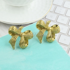May include: A pair of gold-tone bow-shaped earrings. The bows have a textured surface and are made of metal.