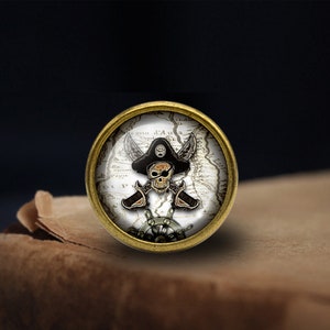 以下が含まれることがあります： ヴィンテージマップを背景に、海賊のドクロとクロスボーンのデザインが施された、金色の丸いボタン。ドクロは黒い海賊の帽子をかぶっています。