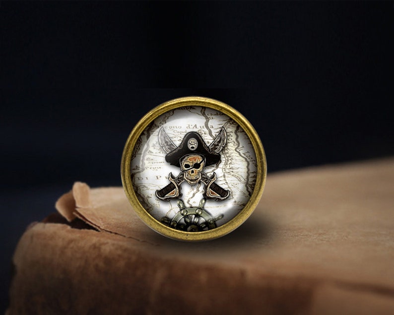 May include: A gold-toned round button with a pirate skull and crossbones design on a vintage map background. The skull is wearing a black pirate hat.