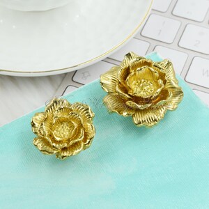May include: Two gold-coloured metal flower-shaped drawer pulls. The larger pull is on the right and the smaller pull is on the left. The pulls are on a light blue surface.