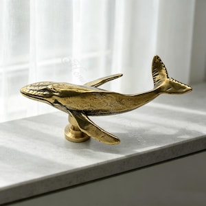 May include: A brass-colored metal whale figurine mounted on a cylindrical pedestal base. The whale is depicted in a horizontal swimming position with its tail fin raised and pectoral fins extended, featuring a textured, weathered metallic finish.