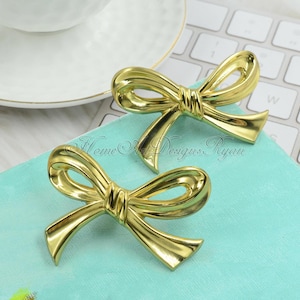 May include: Two shiny gold-colored bow-shaped decorative objects. The bows are positioned on a light blue surface, with a white teacup and a computer keyboard in the background. The bows have a detailed design and a metallic finish.