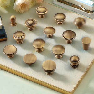 Puede incluir: Una colecci&oacute;n de tiradores y pomos de armario de lat&oacute;n antiguo en varios dise&ntilde;os. Los pomos son redondos, algunos con detalles decorativos como estr&iacute;as y c&iacute;rculos conc&eacute;ntricos. El acabado tiene un aspecto c&aacute;lido y envejecido, lo que sugiere una est&eacute;tica vintage.