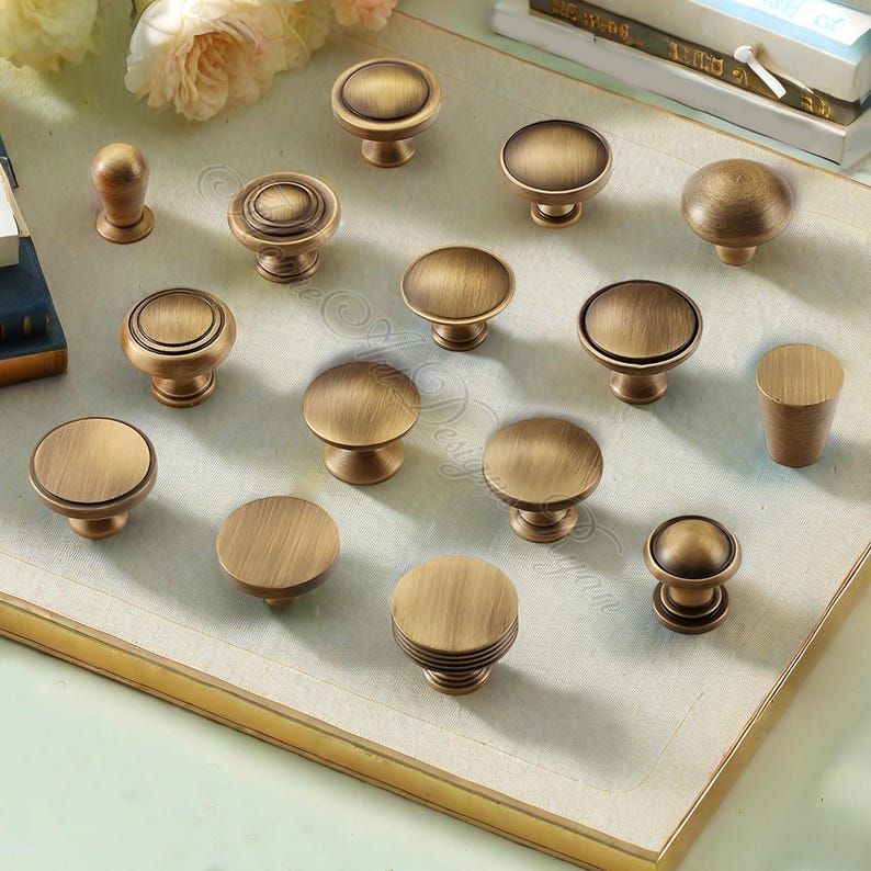 Puede incluir: Una colecci&oacute;n de tiradores y pomos de armario de lat&oacute;n antiguo en varios dise&ntilde;os. Los pomos son redondos, algunos con detalles decorativos como estr&iacute;as y c&iacute;rculos conc&eacute;ntricos. El acabado tiene un aspecto c&aacute;lido y envejecido, lo que sugiere una est&eacute;tica vintage.