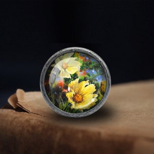 May include: A silver knob with a round glass top featuring a painted image of yellow flowers with a blue and green background.
