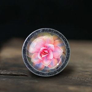 Peut inclure: Un bouton rond de couleur argent avec un insert en verre représentant une fleur de succulente rose et violette.
