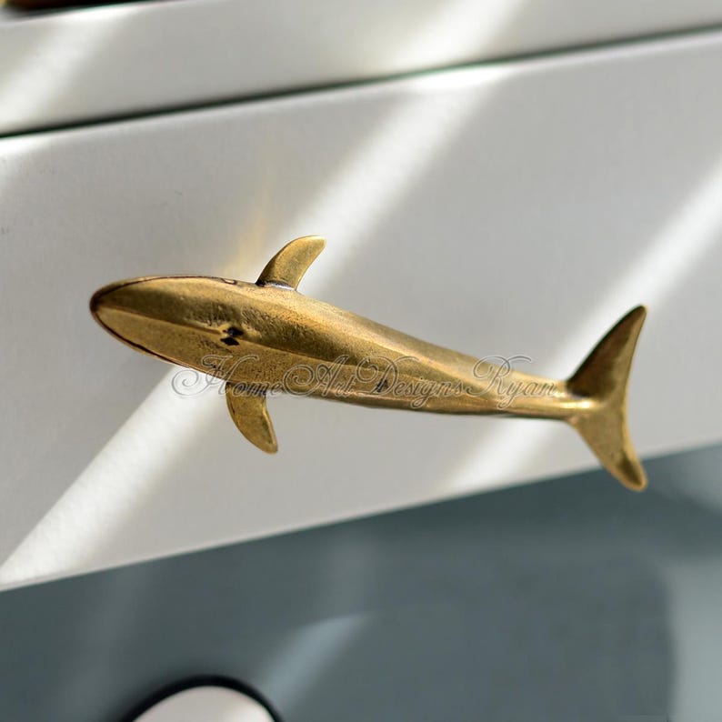 May include: A gold-toned whale-shaped drawer pull. The whale is facing left and has a slightly curved body. The pull is made of metal and has a smooth finish.