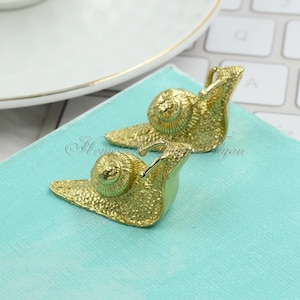 Op de afbeelding: Twee gouden slakkenbeeldjes, een op de ander, met gedetailleerde texturen van de schelp en het lichaam.