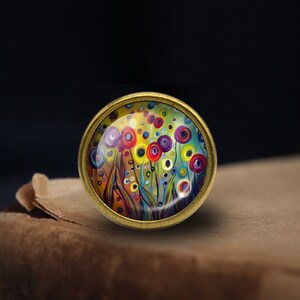 Peut inclure: Un pendentif rond de couleur laiton avec une peinture abstraite colorée de fleurs et de cercles. La peinture est encadrée dans un dôme de verre transparent.