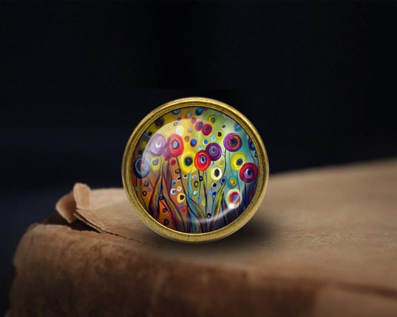 May include: A round, brass-colored pendant with a colorful abstract painting of flowers and circles. The painting is set in a clear glass dome.