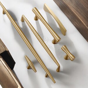 Puede incluir: Una colecci&oacute;n de herrajes dorados para armarios, que incluye varios dise&ntilde;os de tiradores y pomos. Los herrajes est&aacute;n dispuestos sobre una superficie blanca, mostrando diferentes formas y tama&ntilde;os para aplicaciones de mobiliario. Los herrajes son de metal.