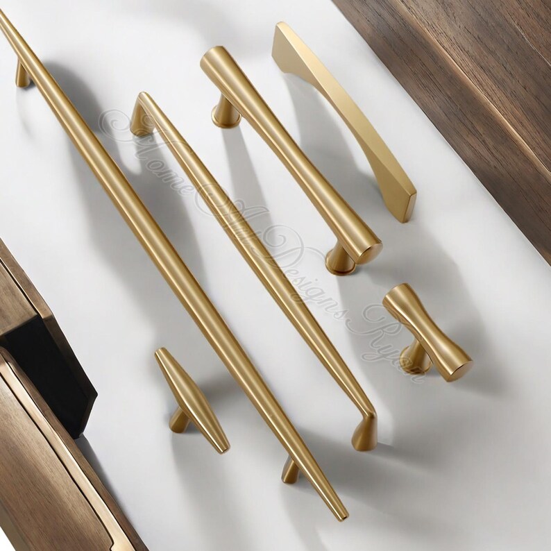 Puede incluir: Una colecci&oacute;n de herrajes dorados para armarios, que incluye varios dise&ntilde;os de tiradores y pomos. Los herrajes est&aacute;n dispuestos sobre una superficie blanca, mostrando diferentes formas y tama&ntilde;os para aplicaciones de mobiliario. Los herrajes son de metal.