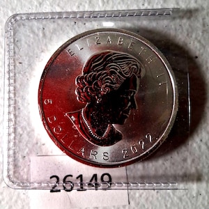 May include: A silver coin featuring a profile of Queen Elizabeth II, with the text "ELIZABETH II" and "5 DOLLARS 2022". The coin is encased in a clear plastic holder, with the number "26149" visible below.