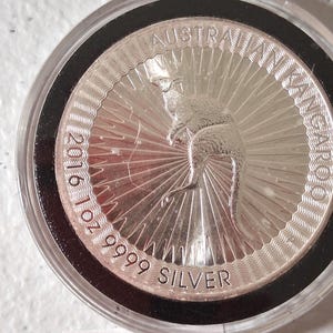 May include: A silver coin featuring a kangaroo, with the text "AUSTRALIAN KANGAROO" and "2016 1 oz 9999 SILVER". The coin is encased in a clear plastic holder with a black border.