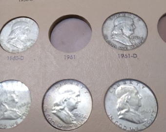 May include: A close-up of a coin collection album with silver coins from 1956 to 1963. The album has a brown leather-like cover and the coins are displayed in individual slots.