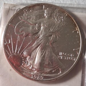 May include: A silver coin featuring a woman draped in flowing robes, holding a branch and shield, with the sun rising behind her. The coin is dated 1989 and includes the inscription "IN GOD WE TRUST".