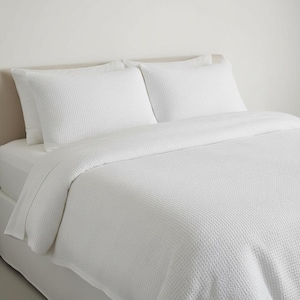 Könnte beinhalten: Ein weißes Bettlaken mit Waffelstruktur und zwei passende Kissenbezüge sind auf einem Bett drapiert. Die Bettwäsche hat eine strukturierte Oberfläche, die ein subtiles Muster erzeugt. Die Gesamtästhetik ist sauber und minimalistisch.