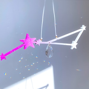 Könnte beinhalten: Ein weiß-rosa Sternenmobil mit einem Kristallprisma, das in der Mitte hängt. Die Sterne sind durch weiße Stäbe verbunden und das Mobile hängt von der Decke.