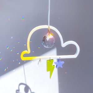 Könnte beinhalten: Ein weiß-gelber Wolken-Mobile mit einem Kristallprisma, das in der Mitte hängt. Das Mobile hat auch einen gelben Blitz, einen weißen Regentropfen und einen blauen Stern, die daran hängen.