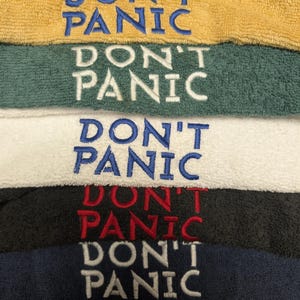 Könnte beinhalten: Ein Stapel bestickter Handtücher in verschiedenen Farben, darunter Gelb, Grün, Weiß, Schwarz und Marineblau. Jedes Handtuch zeigt den gestickten Schriftzug "DON'T PANIC" in kontrastierenden Farben wie Blau, Weiß und Rot.