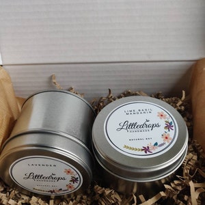 May include: Two round silver metal tins containing natural soy candles. One label reads Lavender, the other Lime, Basil, Mandarin, both featuring the brand name Littledrops Handmade and floral illustrations. The tins are nestled in brown paper filler.