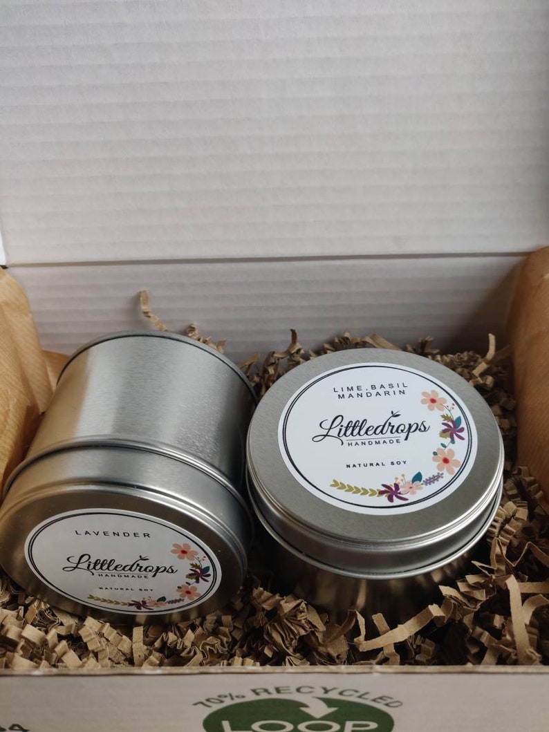 May include: Two round silver metal tins containing natural soy candles. One label reads Lavender, the other Lime, Basil, Mandarin, both featuring the brand name Littledrops Handmade and floral illustrations. The tins are nestled in brown paper filler.