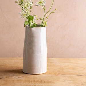 Könnte beinhalten: Ein weißer Keramik Krug mit leicht ausgestelltem Rand, gefüllt mit einem Strauß zarter weißer Blumen mit grünen Stielen. Der Krug steht auf einer hellbraunen Holzoberfläche.