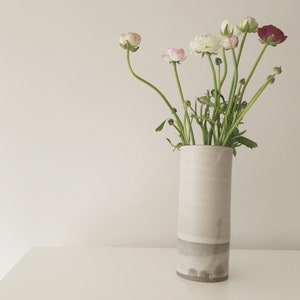 May include: A white ceramic vase with a grey stripe around the middle, filled with white and pink ranunculus flowers.