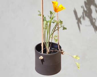 Op de afbeelding: Een zwarte keramische hangende plantenpot met een enkele gele bloem en groene bladeren. De pot wordt opgehangen met twee bruine leren banden.