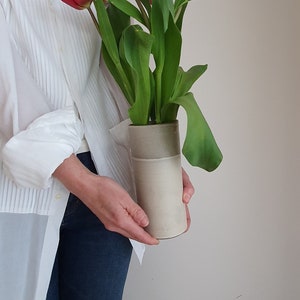 May include: A person holding a white ceramic vase with a bouquet of red tulips.