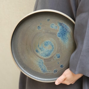 May include: A large, round ceramic plate with a dark grey glaze and a swirling blue design. The plate has a slightly raised rim.