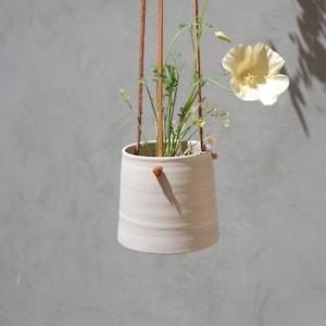 May include: A white ceramic hanging planter with a single yellow flower and green leaves. The planter is suspended by brown leather straps.