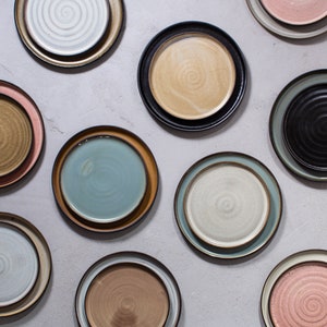 Pode incluir: Uma cole&ccedil;&atilde;o de dez pratos de cer&acirc;mica em v&aacute;rias cores e padr&otilde;es, incluindo azul, verde, marrom e rosa. Os pratos est&atilde;o dispostos em um padr&atilde;o de grade sobre um fundo cinza claro.