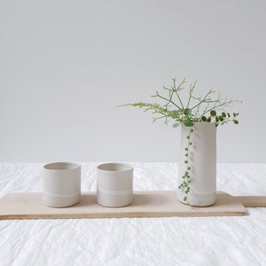 May include: Three white ceramic vases with a single band of white glaze around the middle. The vase on the right is taller and holds a sprig of green foliage. The vases are arranged on a wooden board.