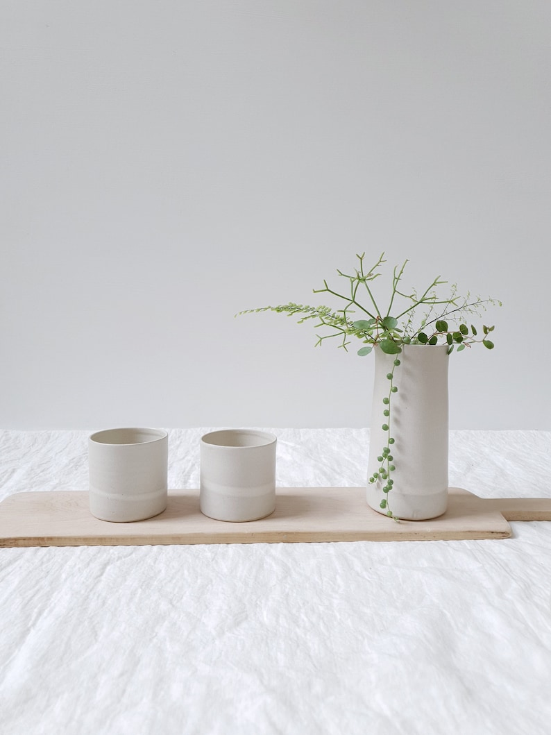 May include: Three white ceramic vases with a single band of white glaze around the middle. The vase on the right is taller and holds a sprig of green foliage. The vases are arranged on a wooden board.
