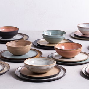 Pode incluir: Um conjunto de tigelas e pratos de cer&acirc;mica em v&aacute;rias cores e padr&otilde;es, incluindo marrom, bege, rosa, azul e branco. As tigelas e pratos est&atilde;o dispostos sobre uma superf&iacute;cie cinza claro.