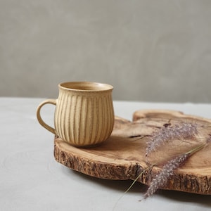 Puede incluir: Una taza de cerámica marrón claro con un diseño acanalado se encuentra sobre una rodaja de madera. La taza tiene una forma redondeada y un asa. La rodaja de madera tiene un borde de corteza natural.