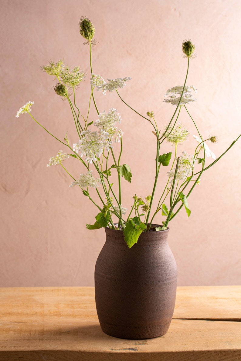 May include: A bouquet of white Queen Anne's Lace flowers in a dark brown ceramic vase on a wooden table.