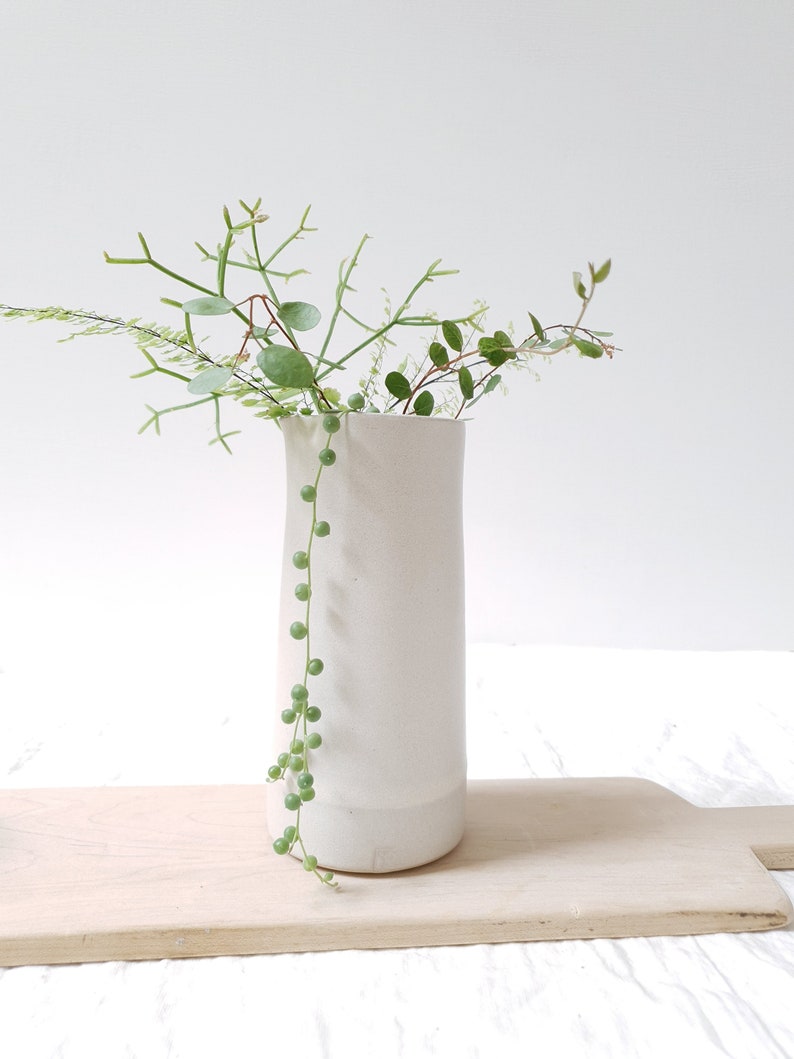 May include: A white ceramic vase with a rounded bottom and a cylindrical shape, filled with green leafy plants.
