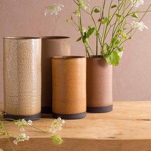 Puede incluir: Cuatro jarrones de cerámica en diferentes colores y texturas, dispuestos sobre una superficie de madera. Los jarrones son todos de forma cilíndrica y tienen un borde marrón oscuro. Un ramo de flores blancas está en el jarrón de la derecha.