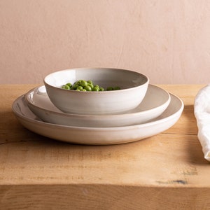 May include: A white ceramic bowl filled with green peas sits on a white ceramic plate, which is on a larger white ceramic plate. The plates are on a wooden table.