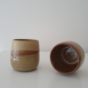 May include: Two ceramic mugs with a brown and beige glaze. The mugs are sitting on a white surface.