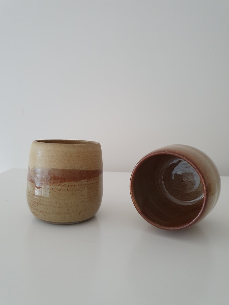 May include: Two ceramic mugs with a brown and beige glaze. The mugs are sitting on a white surface.