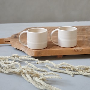 May include: Two white ceramic mugs with rounded handles sit on a wooden cutting board. The mugs are slightly textured and have a subtle off-white color. The cutting board is made of light wood and has a natural grain pattern.