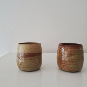 May include: Two ceramic mugs with a brown and tan glaze. The mug on the left has a light brown glaze with a darker brown band around the middle. The mug on the right has a darker brown glaze with a lighter brown band around the middle.