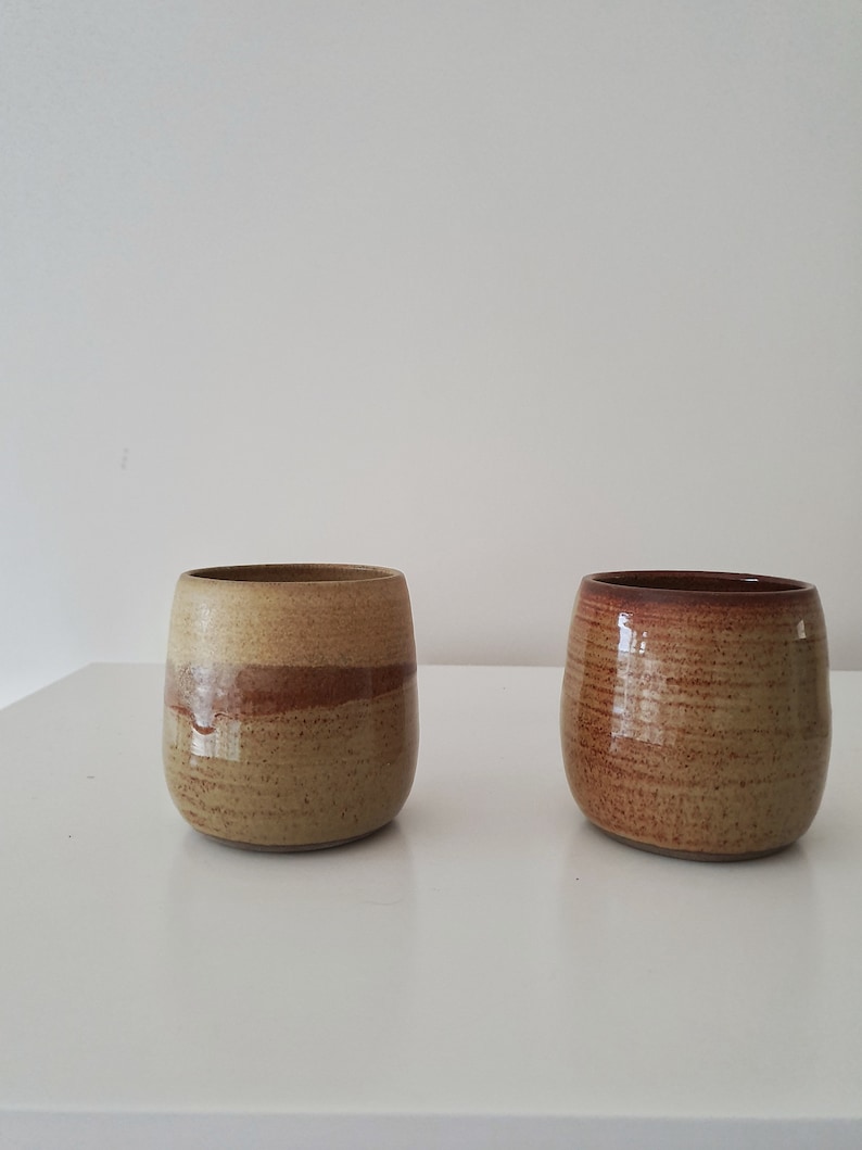 May include: Two ceramic mugs with a brown and tan glaze. The mug on the left has a light brown glaze with a darker brown band around the middle. The mug on the right has a darker brown glaze with a lighter brown band around the middle.
