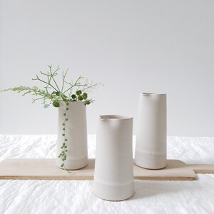 May include: Three white ceramic vases, two with a slightly flared lip and one with a straight lip, on a wooden surface. The vase in the center has a sprig of green foliage.